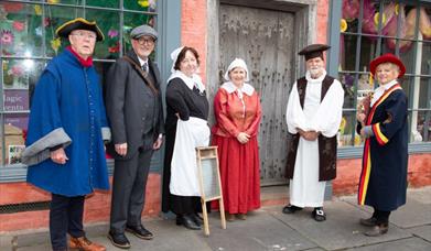 Six tour guides dressed in costumes from different historical eras standing outside a timber framed Tudor house