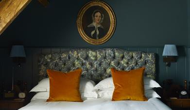 A wooden ceiling beam above a teal-coloured wall with an antique picture of a woman, which hangs above a double bed with patterned headboard and ochre